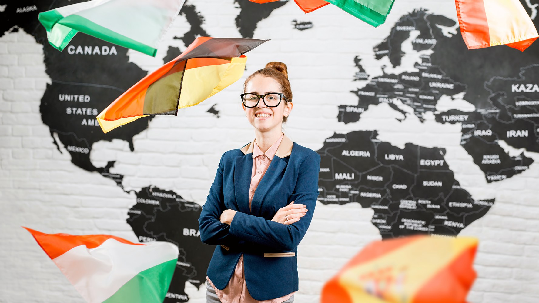 Une jeune enseignante se tient devant la carte du monde accrochée au mur, autour d'elle flottent les drapeaux de différents pays.